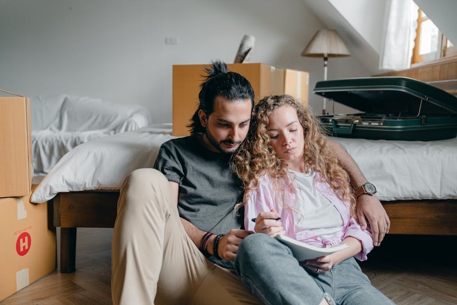A man and woman sit on the floor beside a bed in a bedroom filled with cardboard boxes and packing materials, indicating an ongoing house move. The man, wearing a black t-shirt and beige trousers, has